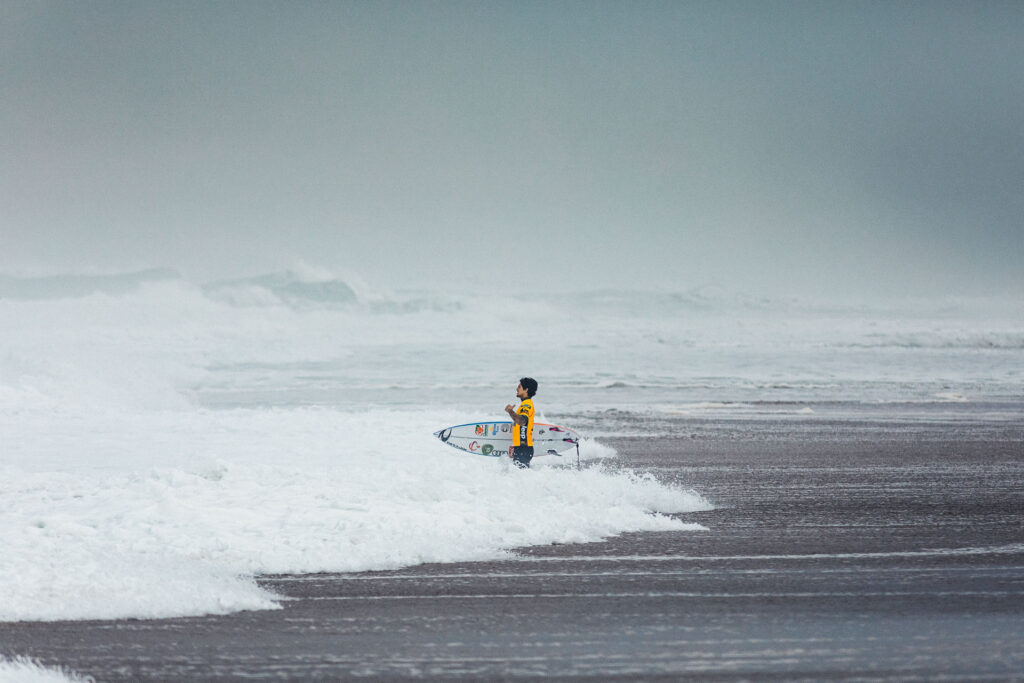Pro surfer Gabriel Medina is standing at the beach large waves in the background by Ondrej Kolacek
